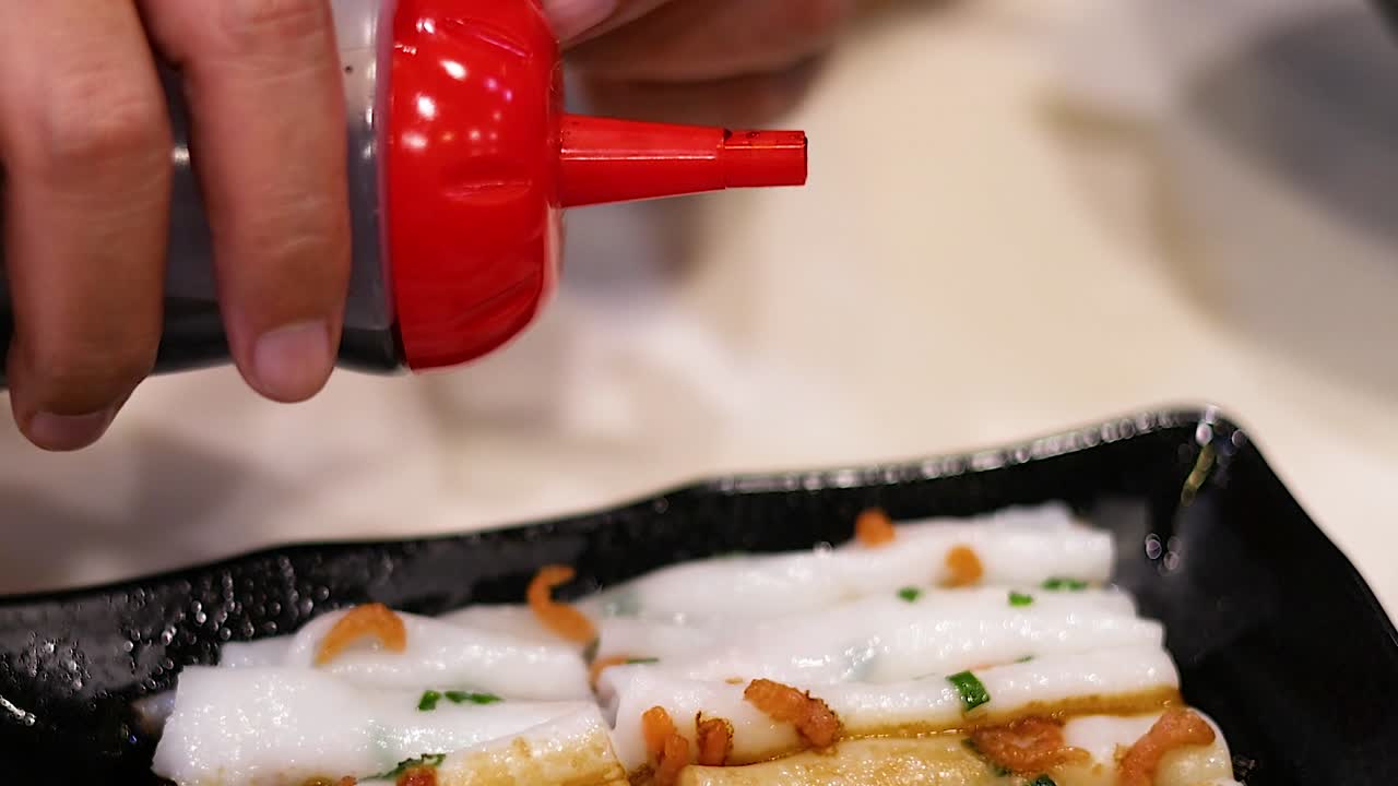 A hand pours dark sauce from a red bottle onto rice noodles served on a black plate.