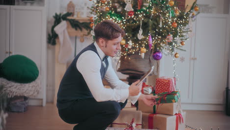 hombre usando una tableta digital para una llamada de video mientras organiza regalos de navidad en casa