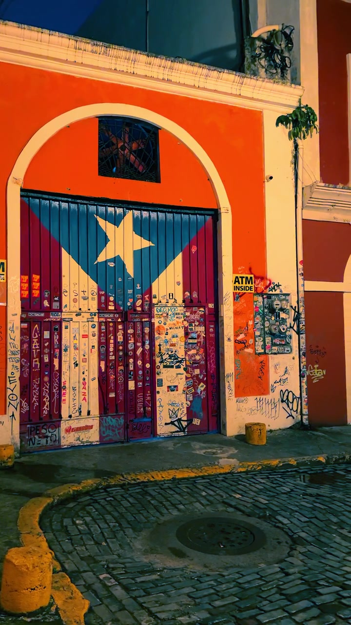 San Juan, Porto Rico, 4 agosto 2025: Bandiera portoricana dipinta su porta di garage di notte. Una grande porta dipinta con il design della bandiera portoricana si trova su una facciata colorata di un edificio su una strada acciottolata di notte.