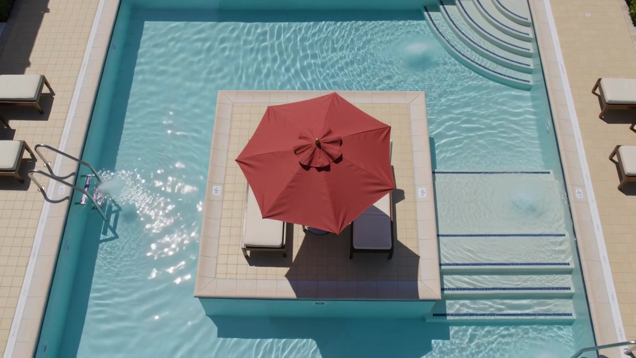 Pulling back drone showing more pool and raised deck with red umbrella, two chairs, water rippling