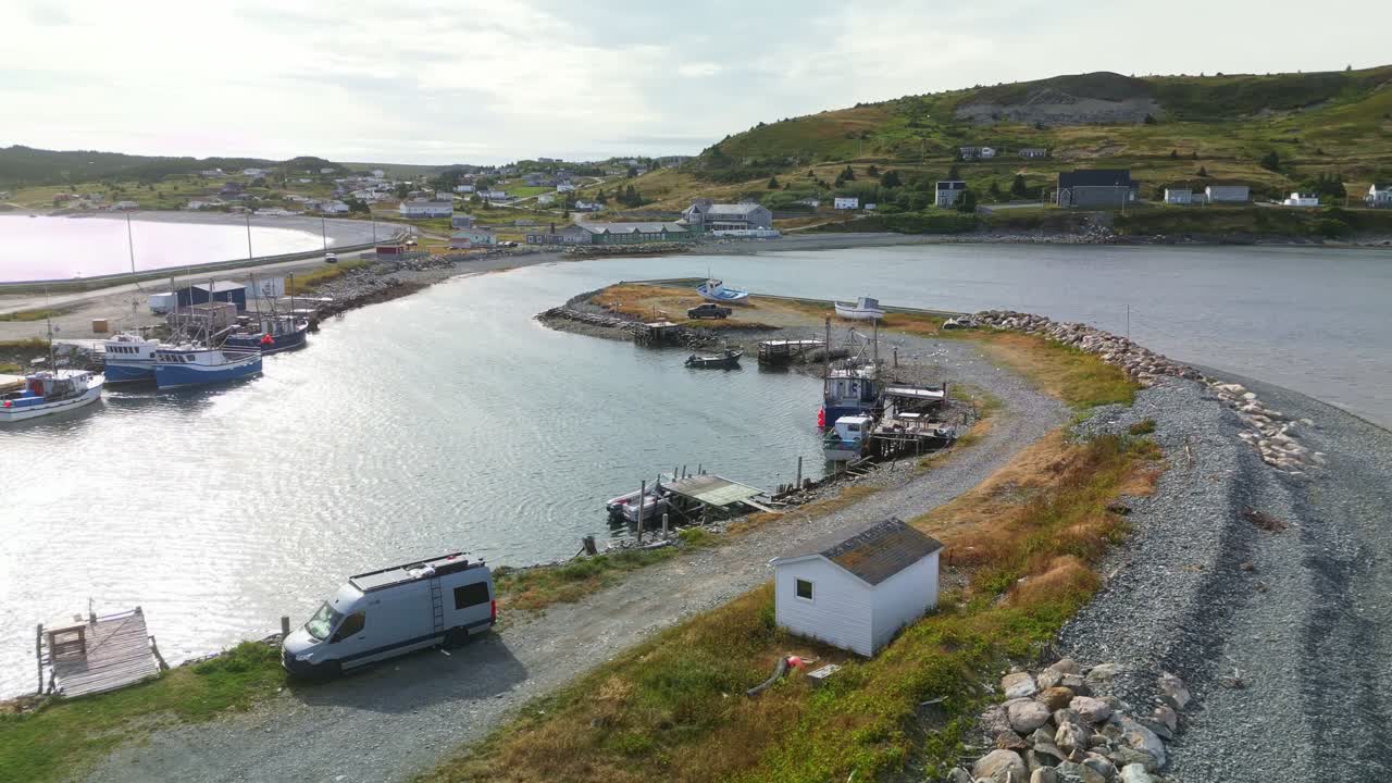 A drone flies over Ferryland’s harbour, capturing a rectangular wharf jutting into calm waters, small boats moored, and coastal hillsides with houses in the distance