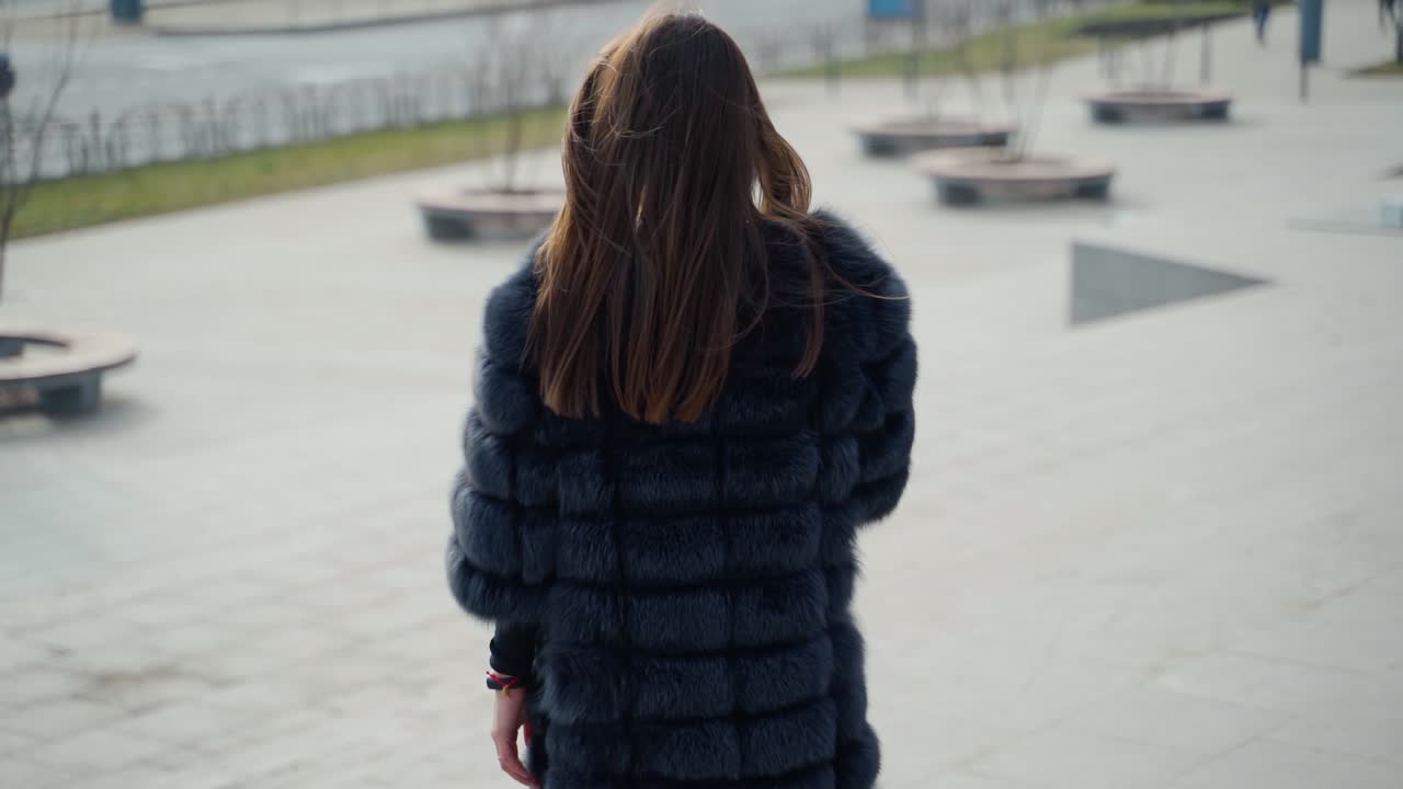 Pensive winter girl in black fur coat outdoors. Back view of a beautiful brunette walking in the city. Attractive luxury woman posing on camera. Slow motion.