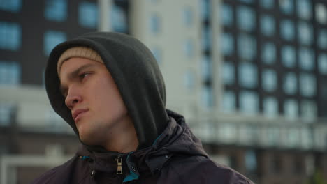 close-up of man turning head thoughtfully while wearing hoodie and beanie, with blurred background featuring modern office buildings