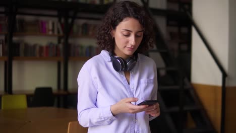 Portrait-Of-Walking-Brunette-Woman-Wearing-White-Blouse,-Looking-On-Smartphone-Screen-In-Her-Hands,-Headphones-On-Neck