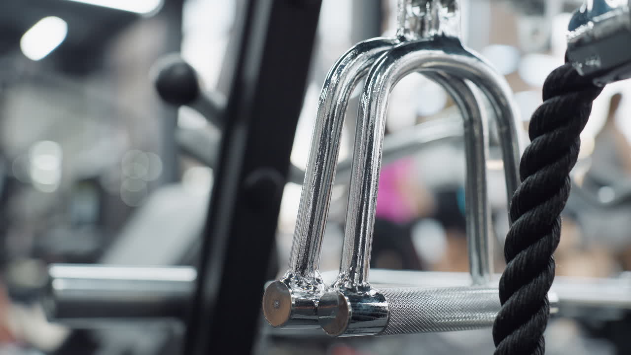 Close up of steel grip handle and rope attachment on cable machine with blurred fitness center background showing faint silhouettes of people exercising and detailed metal texture focus