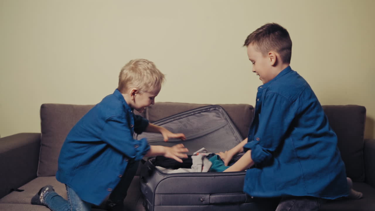 Joyful boys wearing identically are taking out clothes of the packed suitcase. Happy brothers making a mess with clothes on the couch in the room. Slow motion.