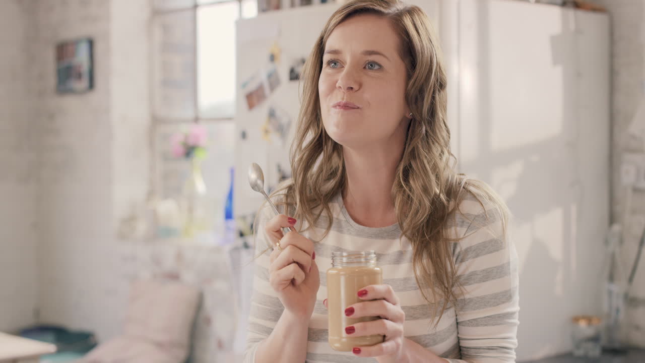 Caucasian girl indulging eating peanut butter from jar using spoon at home in the morning