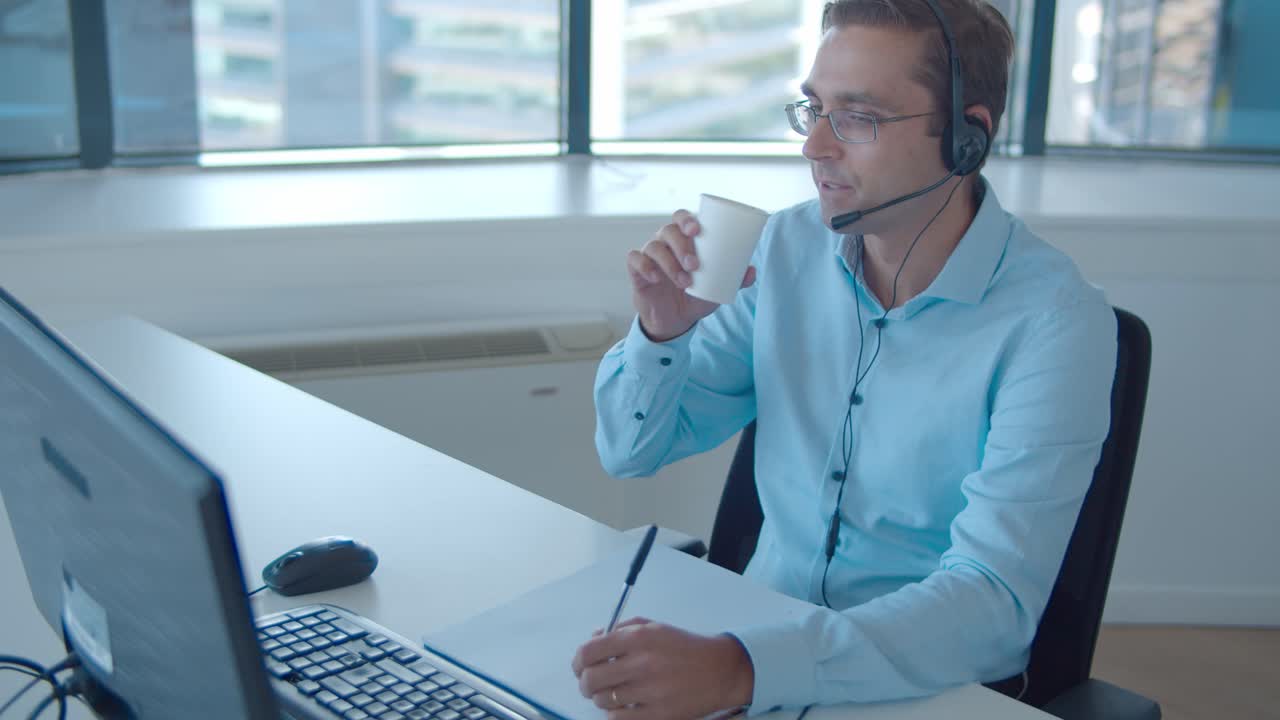 operador masculino positivo en auriculares tomando llamadas y bebiendo agua