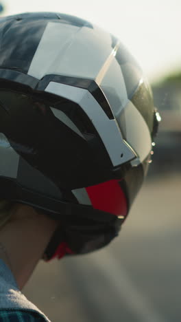 close-up of a woman wearing a helmet, observing cars passing by on a road, a blurred red motorcycle and someone s clothing are visible in the background