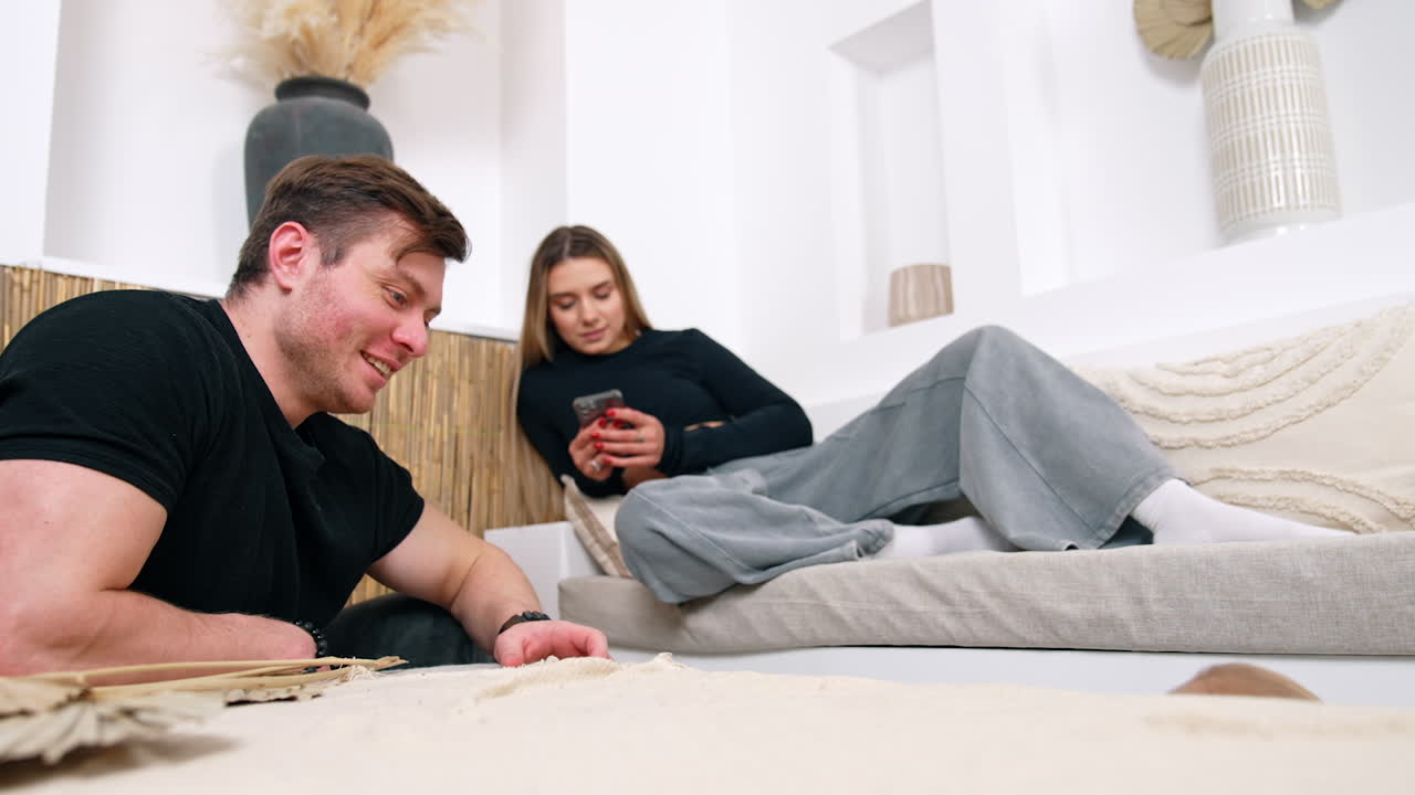 Caucasian man sits on the floor looking after his son. Woman lies on sofa with phone in hands at backdrop.