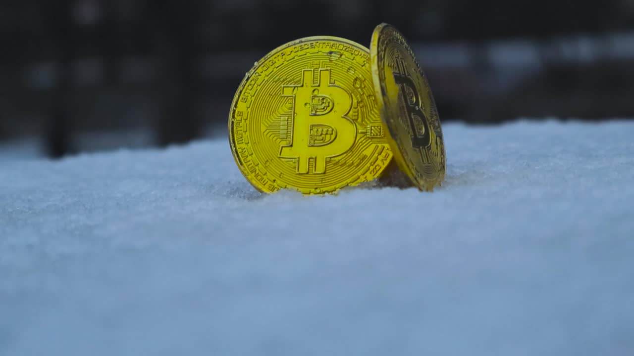 dos monedas de criptomoneda de bitcoin reflectantes de color dorado de pie en la nieve gruesa blanca al aire libre durante el invierno, mientras la nieve cae sobre y alrededor de las monedas.