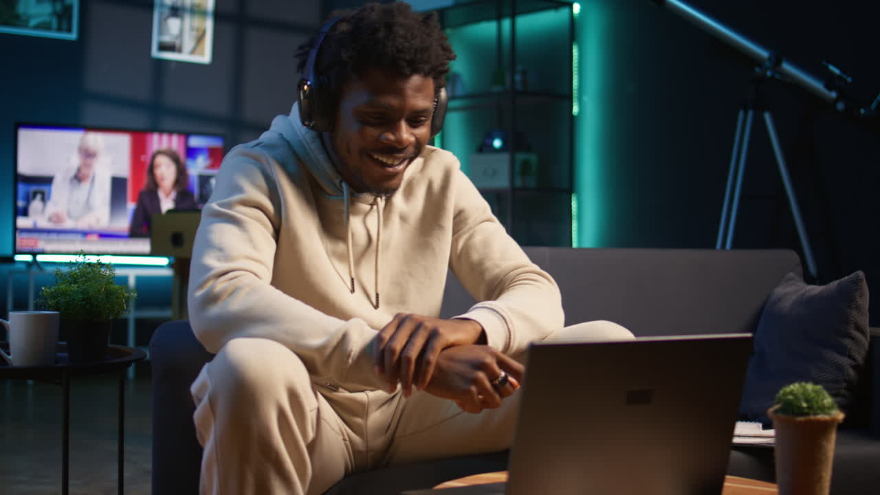 African american male person listening to a podcast and multitasking on the coffee table, browsing the internet and navigating through his laptop with headphones on. Self employed man. Camera B.