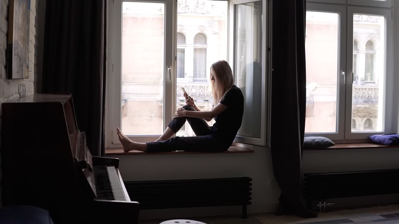 Young woman sitting on window sill and using smartphone in living room with piano