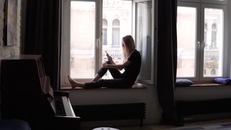 Young-woman-sitting-on-window-sill-and-using-smartphone-in-living-room-with-piano