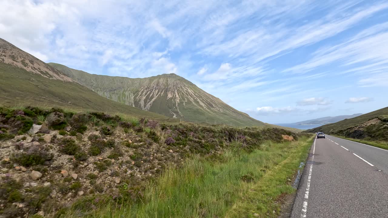 A vehicle travels a winding rural road through lush green hills and mountains under bright daylight, captured in a smooth forward motion with wide landscape views