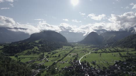 video frontal de drones moviéndose sobre granjas y casas en el triglav con colinas en el horizonte, el sol al frente y cielo despejado con nubes
