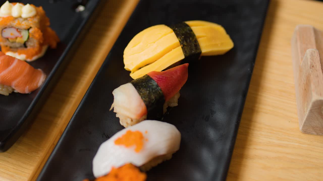 Hand reaches for tamago nigiri sushi on black plate, bright natural lighting, close-up view