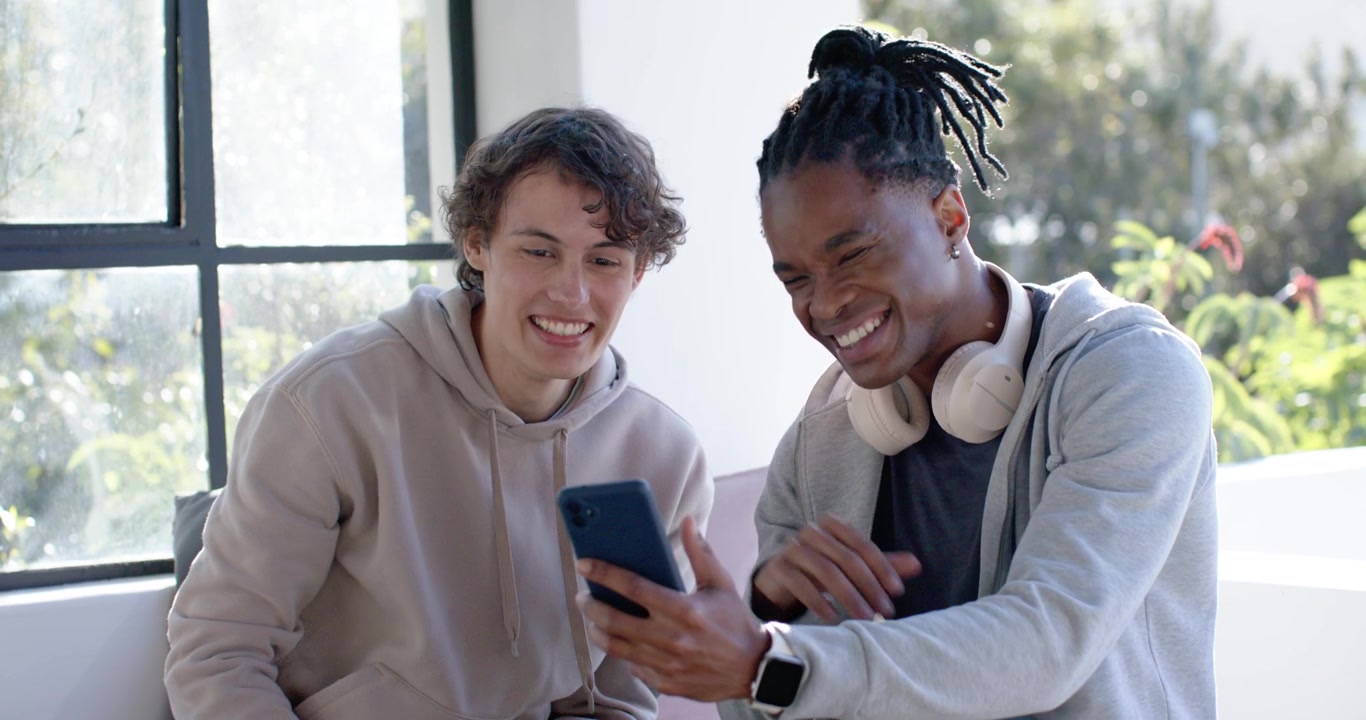 Diverse male friends sitting on couch in lounge; left man pointing at smartphone
