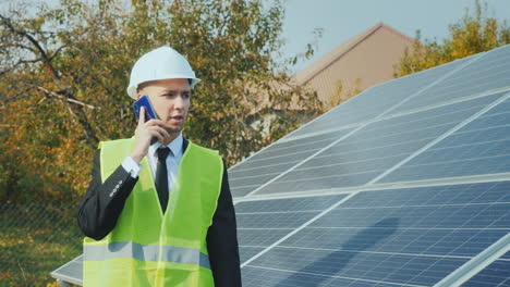 A-Worker-In-A-Helmet-Walks-Along-A-Row-Of-Solar-Panels-Speaks-On-The-Phone