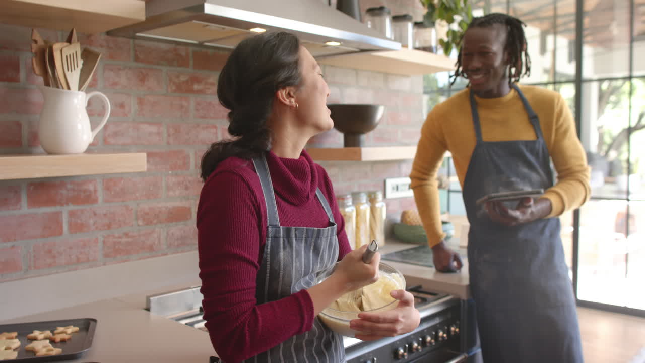 pareja feliz en delantales usando tableta, mezclando masa y hablando en la cocina, cámara lenta