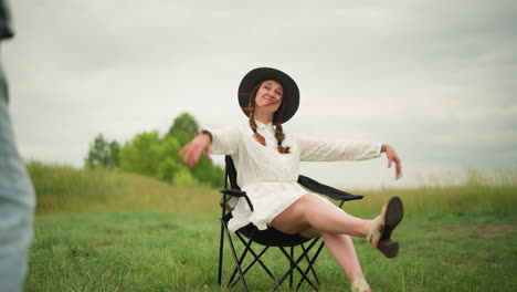 a woman wearing a white dress and a black hat sits on a chair in a grassy field, joyfully waving her hands in the air and posing with a bright smile outdoor scene captures a lively, carefree moment