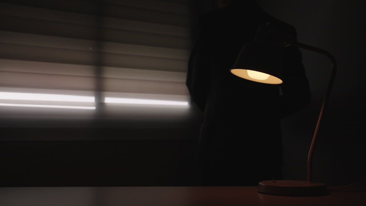 Man lowers window blinds leaving office in darkness with warm desk lamp light. Concept of stress, burnout, overtime work or end of business day