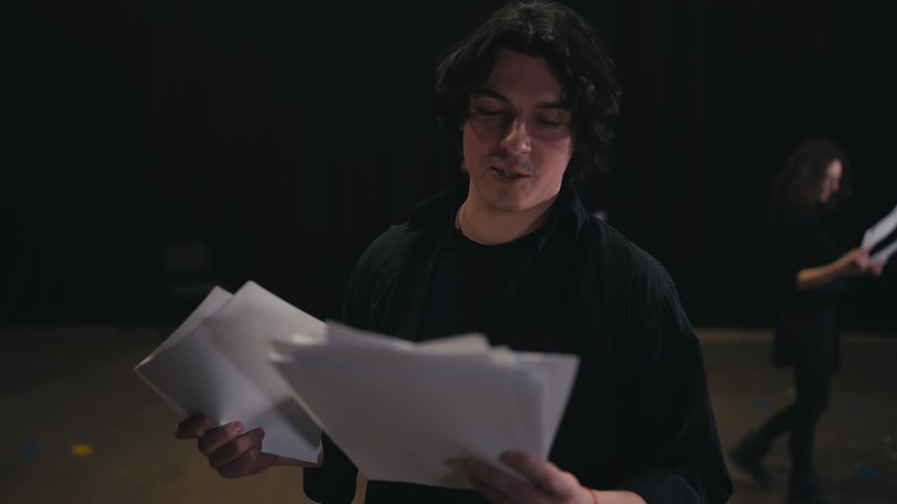 A group of actors are preparing for a performance. A young brunette man in a black suit reads out his words and scripts from sheets of paper during his preparation for a rehearsal for a performance in a theater on stage