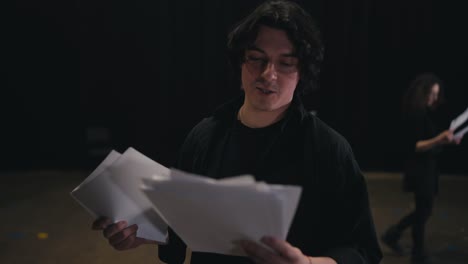 A-group-of-actors-are-preparing-for-a-performance.-A-young-brunette-man-in-a-black-suit-reads-out-his-words-and-scripts-from-sheets-of-paper-during-his-preparation-for-a-rehearsal-for-a-performance-in-a-theater-on-stage