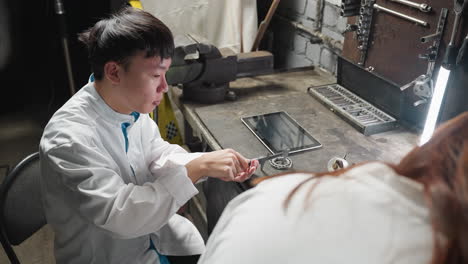 close-up back view of observer watching a young technician in a white lab coat working on a tablet and picking up precision equipment in an automotive workshop, illuminated by lamp light