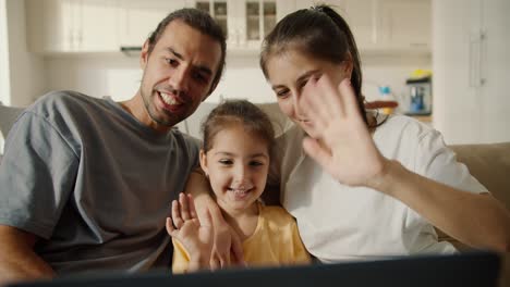 Happy-family,-a-brunette-guy-with-stubble,-his-brunette-girlfriend-in-a-white-T-shirt-and-their-joyful-daughter-in-a-yellow-dress-communicate-with-their-family-and-friends-via-video-conference-using-a-laptop-in-a-modern-studio-room