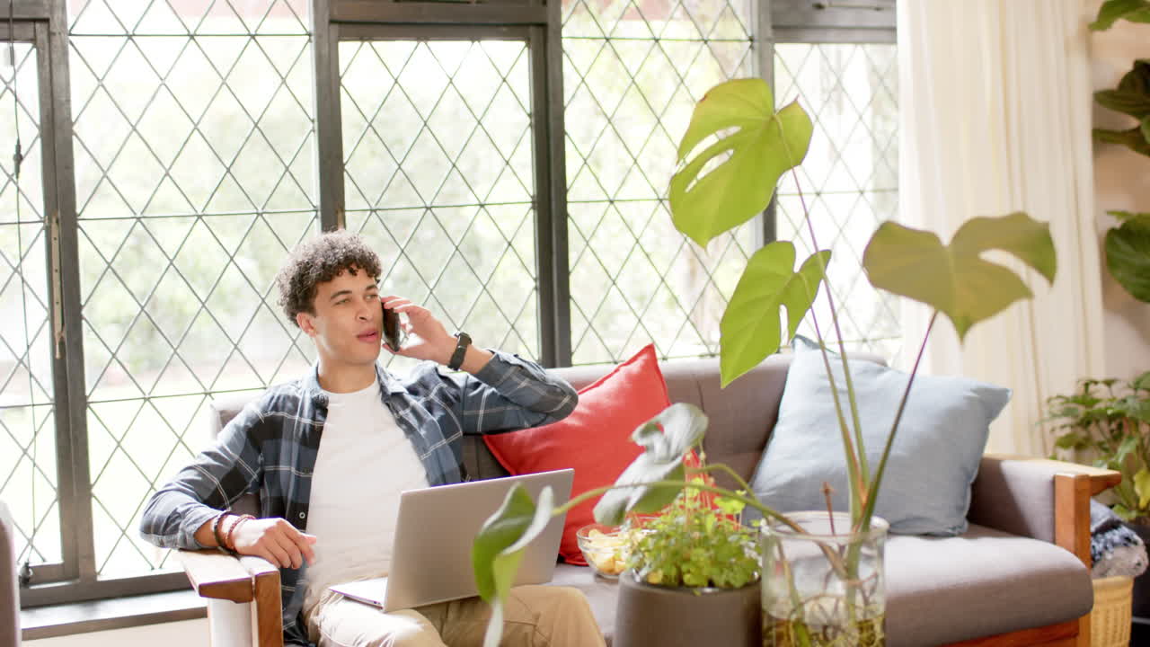 hombre biracial trabajando desde casa usando portátil y hablando en teléfono inteligente, cámara lenta