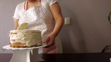 Mujer-Latina-Con-Un-Delantal-Preparando-La-Cocina-Horneando-Un-Pastel-Esparciendo-Glaseado-De-Mantequilla-Con-Un-Raspador-De-Pastel-Blanco