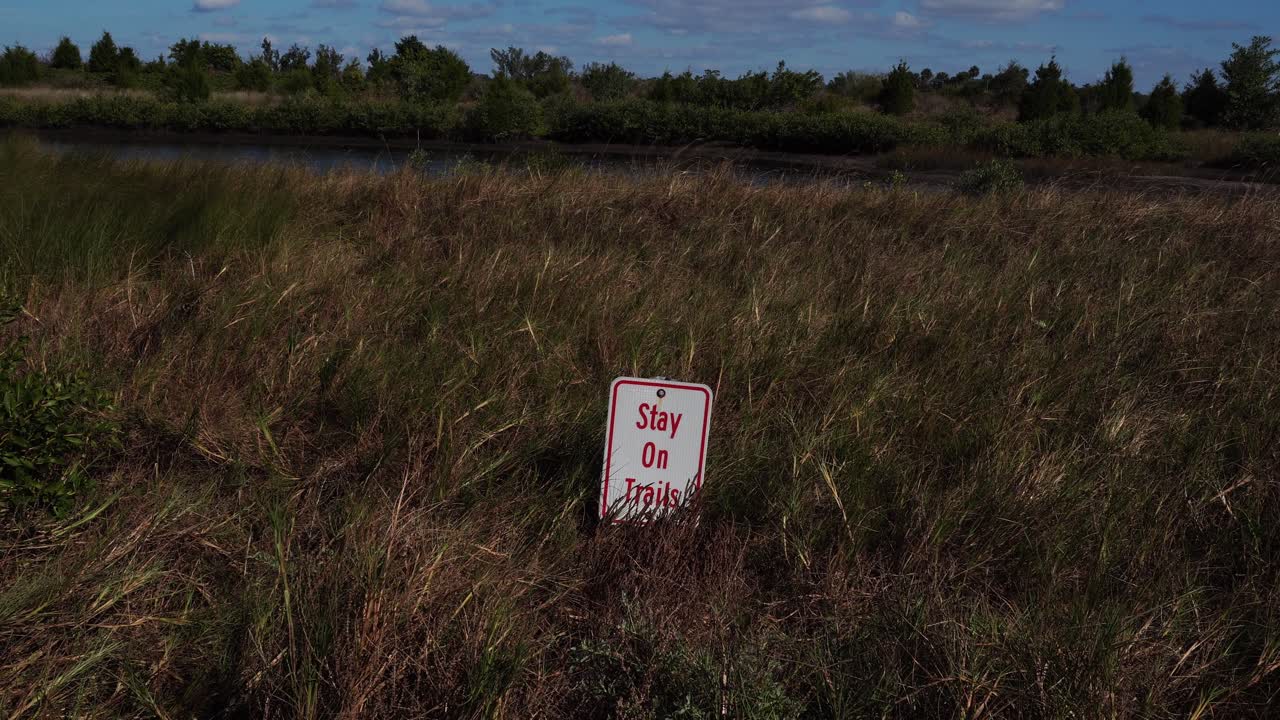 Um campo com vegetação muito baixa possui uma placa indicando para permanecer nas trilhas. Ao fundo, pode-se ver água, árvores e um céu com nuvens