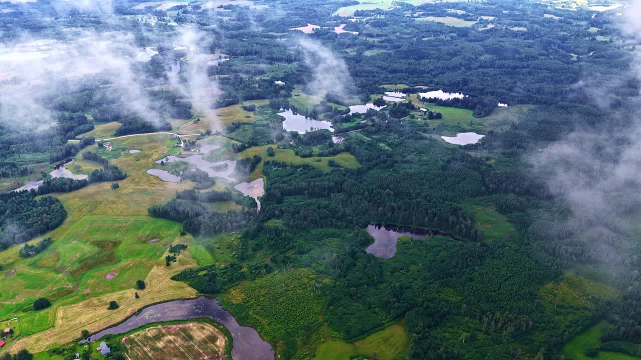 A majestic aerial panning shot reveals the tranquil and picturesque Latvian countryside, a peaceful blend of dense forests, agricultural fields, winding roads, and pristine lakes