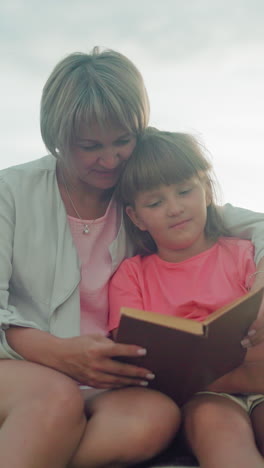 hermosa escena de una madre sosteniendo a su hija cerca, ambos sentados en el heno con un sombrero cercano, en un campo abierto al anochecer, compartiendo un libro