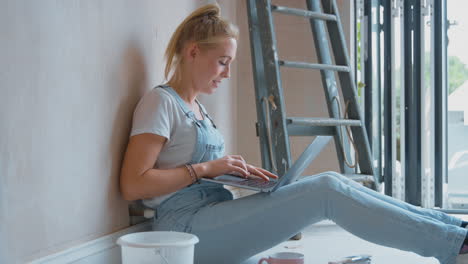 woman renovating room at home sits on floor with laptop taking a break from decorating