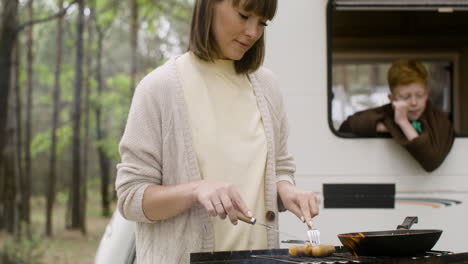 woman cooking sausages on barbecue grill while her son looking out from campervan window