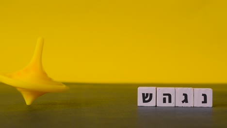 Spinning-yellow-top-Hanukkah-careers-about-beside-the-four-hebrew-letters-nun,-gimel,-heh-and-shin-shown-in-a-static-shot-against-a-two-tone-background