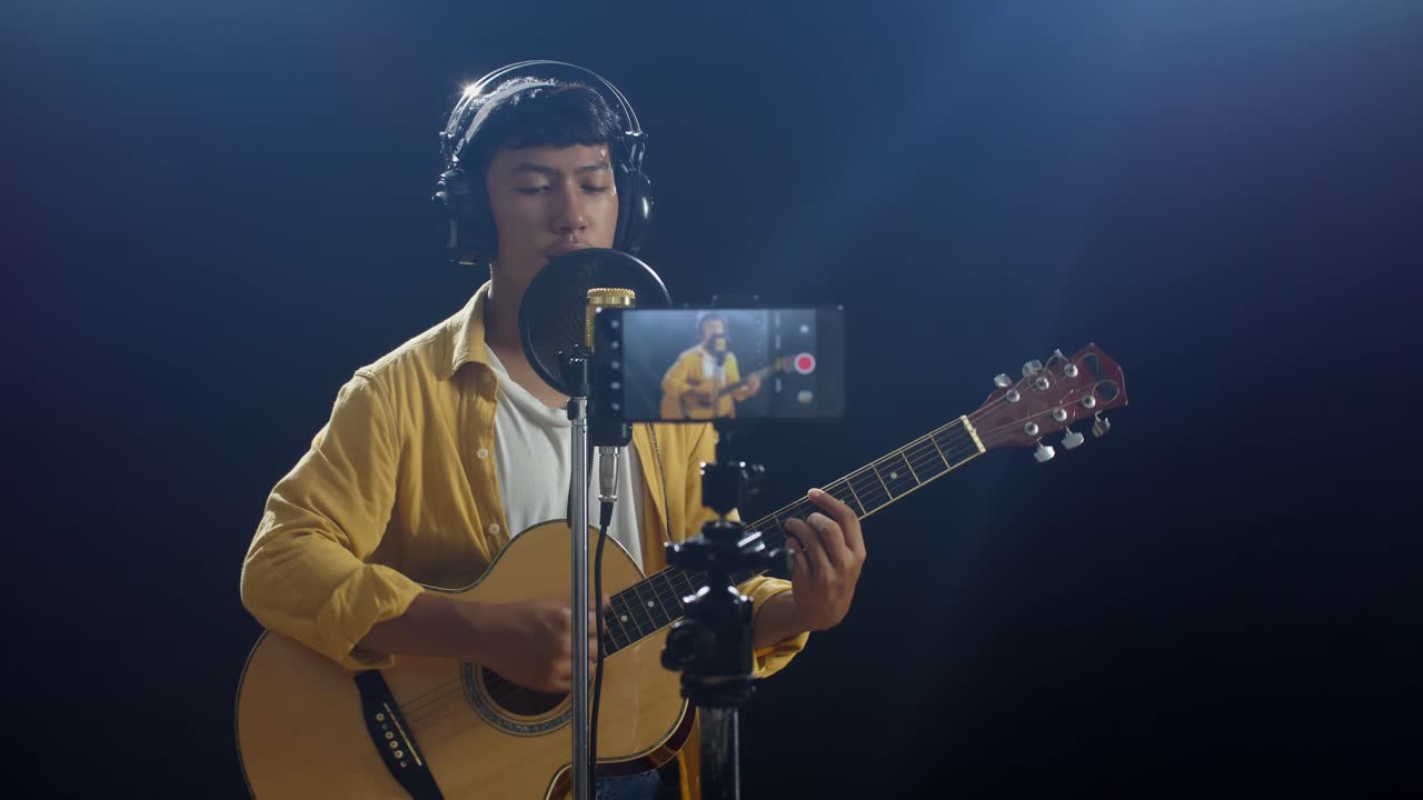 흑색 배경 에 스마트폰 으로 비디오 를 녹음 하는 동안 기타 를 연주 하고 콘덴서 마이크 로 노래 하는 헤드폰 을 착용 한 젊은 아시아 소년