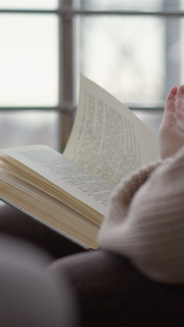 vista lateral de cerca de una joven leyendo un libro, con un fondo de invierno borroso, parece centrada y relajada en un ambiente interior acogedor, con nieve visible fuera de la ventana