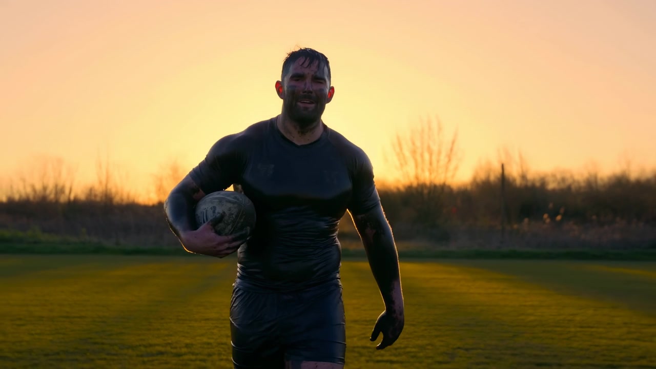 Jugador de rugby masculino camina hacia la cámara a través de un campo de hierba al atardecer, con un balón manchado de barro bajo el brazo, con luz de contorno cálida, silueta oscura y una energía atlética cruda, con rugby, jugador, masculino.