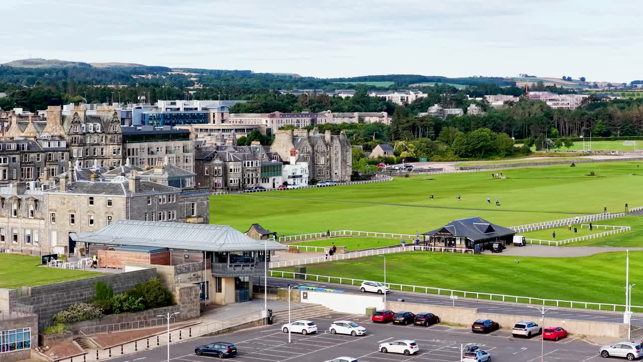 Daytime aerial pan reveals golfers, putting green, historic clubhouse, and townscape at St Andrews