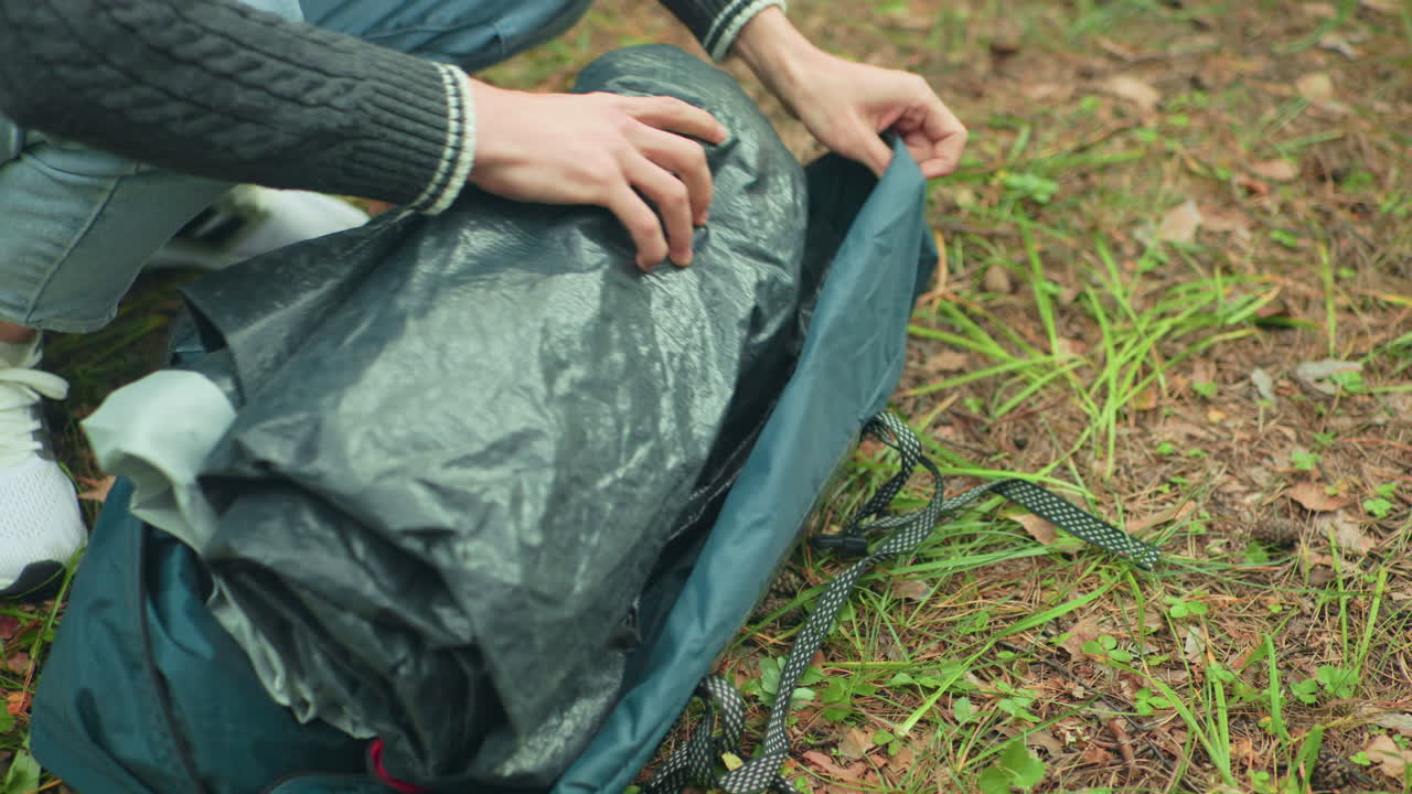 close up of camper with fair skin packing black folded tent into green duffel bag while kneeling on forest floor, surrounded by grass and dry leaves during outdoor camping activity in woodland