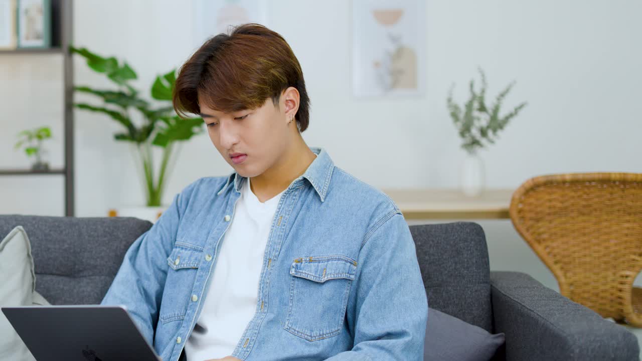 デニムシャツを着た不満そうな若い男性が、自宅でノートパソコンを使ってリモートワークをしている。