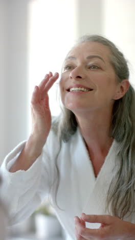 vertical video: applying skincare, woman in bathrobe smiling and looking in mirror at home