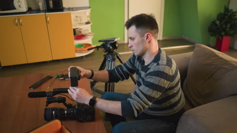 person seated on sofa in home studio, reaching into camera bag to retrieve gear like microphone and lens. preparing for video shoot with camera setup, focused on equipment