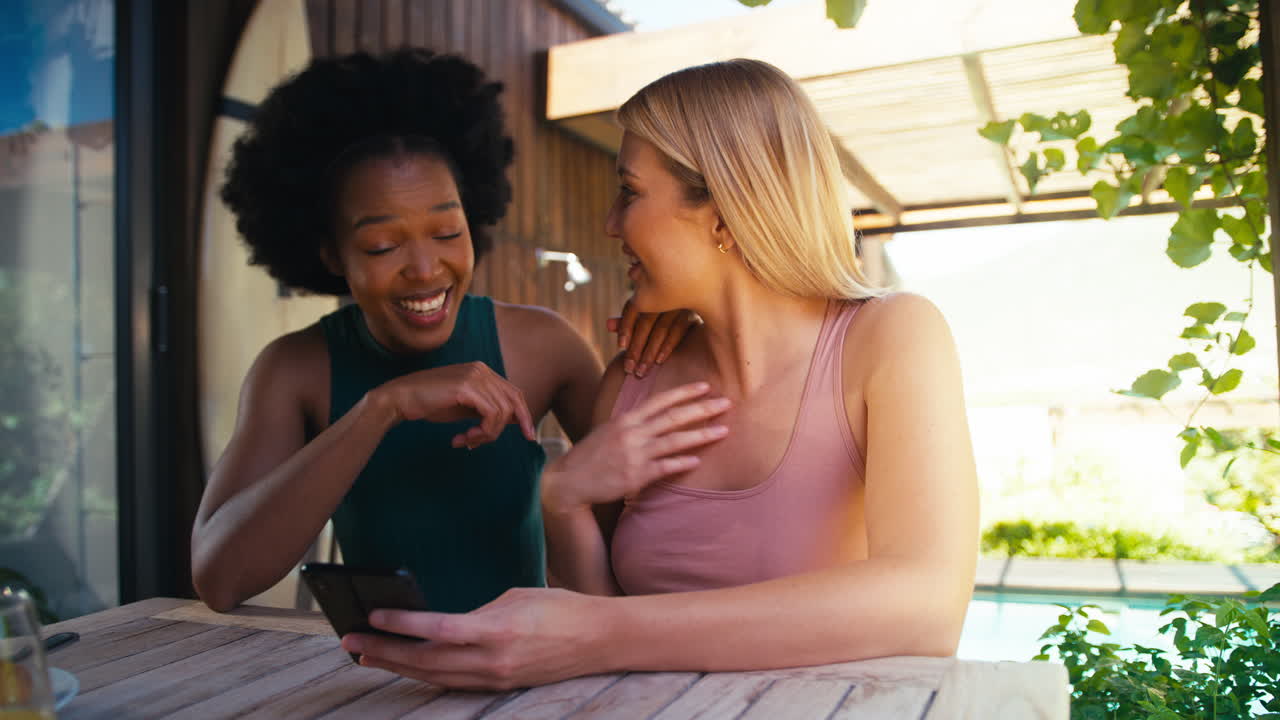 Two Smiling Multi-Cultural Female Friends Outdoors At Home Looking At Mobile Phone
