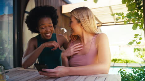 two smiling multi-cultural female friends outdoors at home looking at mobile phone