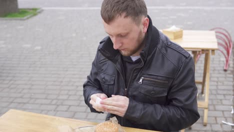 Young-man-sitting-in-a-street-cafe-and-cleaning-his-hands-with-a-napkin-before-taking-tasty-burger-with-french-fries-on-the