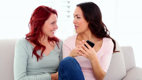 encantadoras mujeres felices llamando emocionadas con un teléfono inteligente sentadas en el sofá